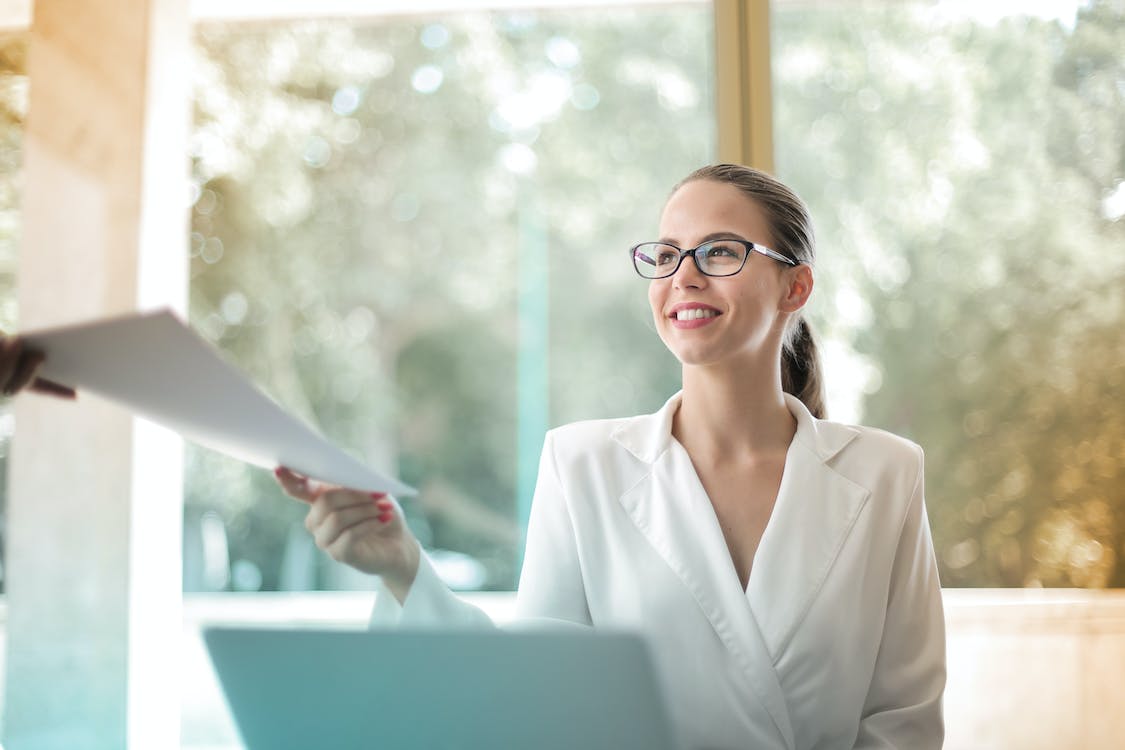 Free Low angle of successful female executive manager in classy style sitting at table with laptop in contemporary workplace and passing documents to colleague Stock Photo